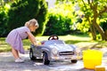 Cute gorgeous toddler girl washing big old toy car in summer garden, outdoors. Happy healthy little child cleaning car Royalty Free Stock Photo