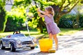 Cute gorgeous toddler girl washing big old toy car in summer garden, outdoors. Happy healthy little child cleaning car Royalty Free Stock Photo