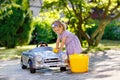 Cute gorgeous toddler girl washing big old toy car in summer garden, outdoors. Happy healthy little child cleaning car Royalty Free Stock Photo