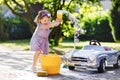 Cute gorgeous toddler girl washing big old toy car in summer garden, outdoors. Happy healthy little child cleaning car Royalty Free Stock Photo