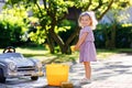 Cute gorgeous toddler girl washing big old toy car in summer garden, outdoors. Happy healthy little child cleaning car