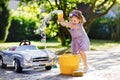 Cute gorgeous toddler girl washing big old toy car in summer garden, outdoors. Happy healthy little child cleaning car Royalty Free Stock Photo