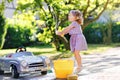 Cute gorgeous toddler girl washing big old toy car in summer garden, outdoors. Happy healthy little child cleaning car Royalty Free Stock Photo