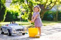 Cute gorgeous toddler girl washing big old toy car in summer garden, outdoors. Happy healthy little child cleaning car Royalty Free Stock Photo