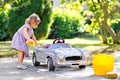 Cute gorgeous toddler girl washing big old toy car in summer garden, outdoors. Happy healthy little child cleaning car Royalty Free Stock Photo