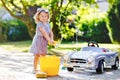 Cute gorgeous toddler girl washing big old toy car in summer garden, outdoors. Happy healthy little child cleaning car Royalty Free Stock Photo