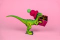 green plastic toy dinosaur wearing knitted hat and holding present box pink background