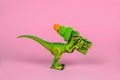 green plastic toy dinosaur wearing knitted hat and holding present box on a pink background