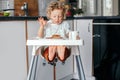 Cute happy Caucasian curly kid boy sitting in high chair eating homemade apple pie and drinking juice. Toddler enjoying food.