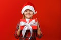 Cute happy child holds a box with a gift. Charming little girl in santa hat and dress on a red background. Royalty Free Stock Photo