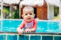 A cute and happy little baby holds to the side of the tropical pool and looks to the camera and laught. Infant girl in swim suit Royalty Free Stock Photo