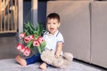 Cute happy little boy holding bouquet of red tulips and soft teddy bear sitting on carpet greeting mother or sister or grandmother