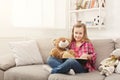 Happy little female child hugging her teddy bear and reading book on sofa at home Royalty Free Stock Photo