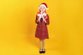 Cute happy little girl holding a box with a christmas gift. Full-length child in a red dress and santa hat on a yellow background