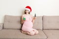 Cute happy pregnant woman in dress at home in santa hat. Mother to be with big belly, hands over tummy. Merry Christmas