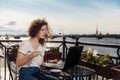 Cute happy young girl working on computer and smiling. Beautiful woman with laptop on a balcony with a landscape on the river Neva Royalty Free Stock Photo