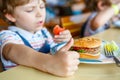 Cute healthy preschool boy eats hamburger sitting in school canteen Royalty Free Stock Photo