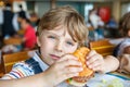 Cute healthy preschool boy eats hamburger sitting in school canteen Royalty Free Stock Photo