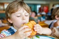 Cute healthy preschool boy eats hamburger sitting in school canteen Royalty Free Stock Photo