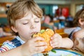 Cute healthy preschool boy eats hamburger sitting in school canteen