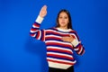 Cute Hispanic girl wearing striped sweater against blue wall. Swearing with the hand on the chest and the open palm