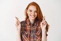 Cute hopeful girl with ginger hair and freckles making a wish, smiling and standing with crossed fingers for good luck