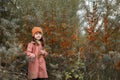 Cute horizontal autumn photo. Serious preschooler girl in an orange hat and a brown coat holds branches of sea buckthorn. in the