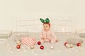 Cute infant girl sitting on Christmas decorations bed and wearing striped long sleeve baby sleeper and festive deer horns, looking