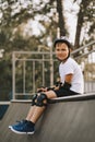 Cute kid boy child in a helmet sitting in a special area in skatepark and holding skateboard. Summer sport activity concept. Happy Royalty Free Stock Photo