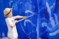 Cute kid boy in straw hat painting fence with blue color. Child drawing outdoors