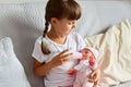 Cute kid at home sitting on sofa. Little girl with pigtails in nursery school, preschooler female child playing with doll, feeding Royalty Free Stock Photo