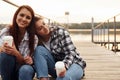 Cute lesbian couple sits together near the lake with cups of drink in hands and embracing each other