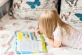 Cute liitle blonde girl lying on a bed and making hometasks in the workbook with a pencil in a hand