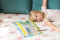 Cute liitle blonde girl lying on a bed and making homework in the workbook with a pencil