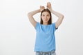 Cute like little bunny. Playful charming adult woman in blue t-shirt, holding hands on head and mimicking animal ears