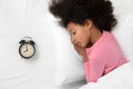 Cute little African-American girl with alarm clock sleeping in bed