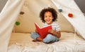 Cute little african american kid curly boy reading book in play tent Royalty Free Stock Photo