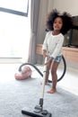 Cute little African girl child with black curly hair vacuuming the floor at bedroom with vacuum cleaner, kid help parents to do