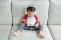 Cute little Asian baby boy in headphones is using a smartphone lying on the sofa at home. Child listening to music on earphones Royalty Free Stock Photo