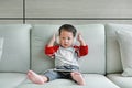 Cute little Asian baby boy in headphones is using a smartphone lying on the sofa at home with looking at camera. Child listening Royalty Free Stock Photo