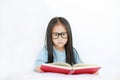 Cute little Asian child girl wearing glasses reading hardcover book lying on bed against white background Royalty Free Stock Photo