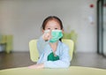 Cute little Asian child girl wearing a protective mask with pointing at you sitting on kid chair in children room Royalty Free Stock Photo