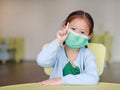 Cute little Asian child girl wearing a protective mask with showing one forefinger sitting on kid chair in children room Royalty Free Stock Photo