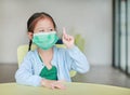 Cute little Asian child girl wearing a protective mask with showing one forefinger sitting on kid chair in children room Royalty Free Stock Photo