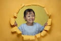 Cute little Asian girl happy smiling poses through torn yellow paper hole, looking something aside Royalty Free Stock Photo