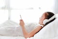 Cute little Asian girl in headphones listen music by smartphone smiling and lying on bed in bedroom at home Royalty Free Stock Photo