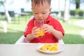 Asian toddler baby boy child sitting in high chair holding & drinking tasty orange juice / cold drink in a Royalty Free Stock Photo