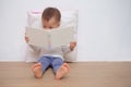 Cute little Asiantoddler boy child sitting on floor, leaning against pillow, looking at a book near white wall, Language developme