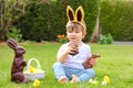 Cute Little Baby Boy With Bunny Ears Eating Chocolate Easter Bunnies Sitting On Green Grass Outside In The Spring Garden With Bask