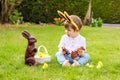 Cute Little Baby Boy With Bunny Ears Eating Chocolate Easter Bunnies Sitting On Green Grass Outside In The Spring Garden